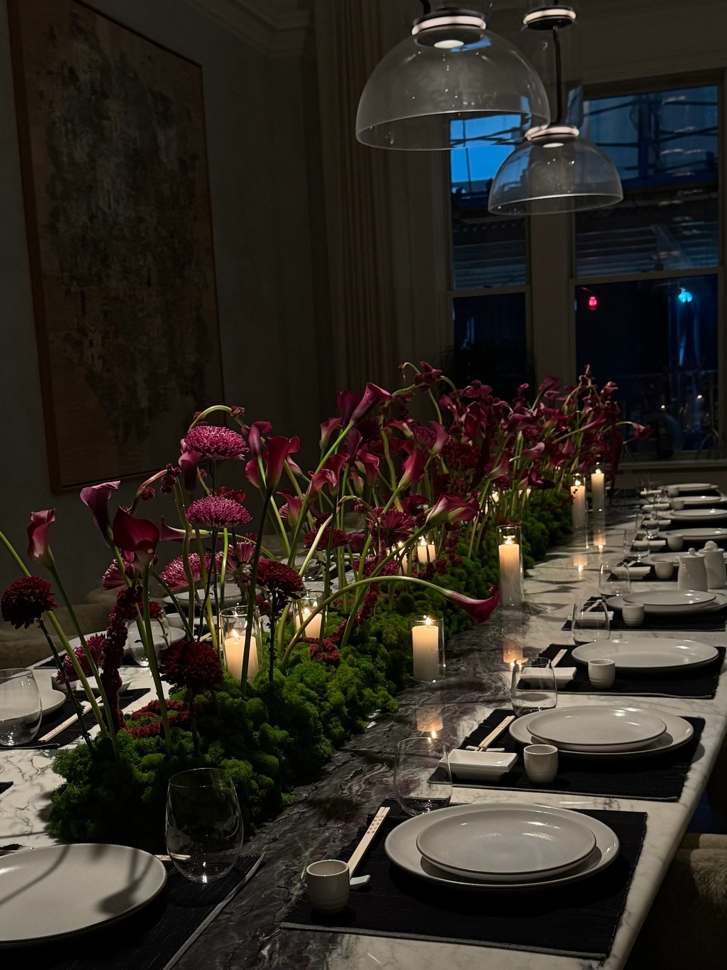 Candlelit dinner table with a moss runner and burgundy calla lilies, set against a downtown New York window at dusk — by Kuration Studio