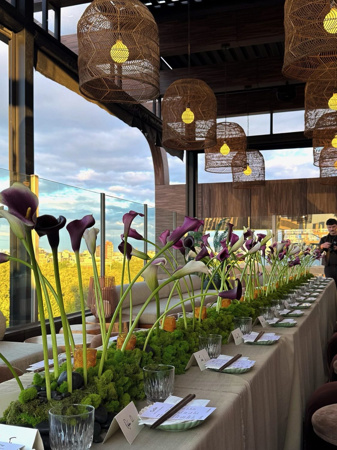 Rooftop dinner at golden hour with calla lilies rising from a moss runner above a city skyline